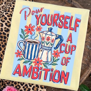 May include: A pale yellow t-shirt featuring a graphic print of a striped coffee mug and a moka pot surrounded by red flowers. The text reads "POUR YOURSELF A CUP OF AMBITION" in bold red letters over a background of light blue vertical stripes.