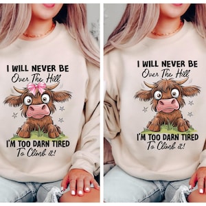May include: A beige sweatshirt featuring a cartoon Highland cow with the text "I WILL NEVER BE Over The Hill I'M TOO DARN TIRED To Climb it!". The cow is brown with a pink bow and star accents.