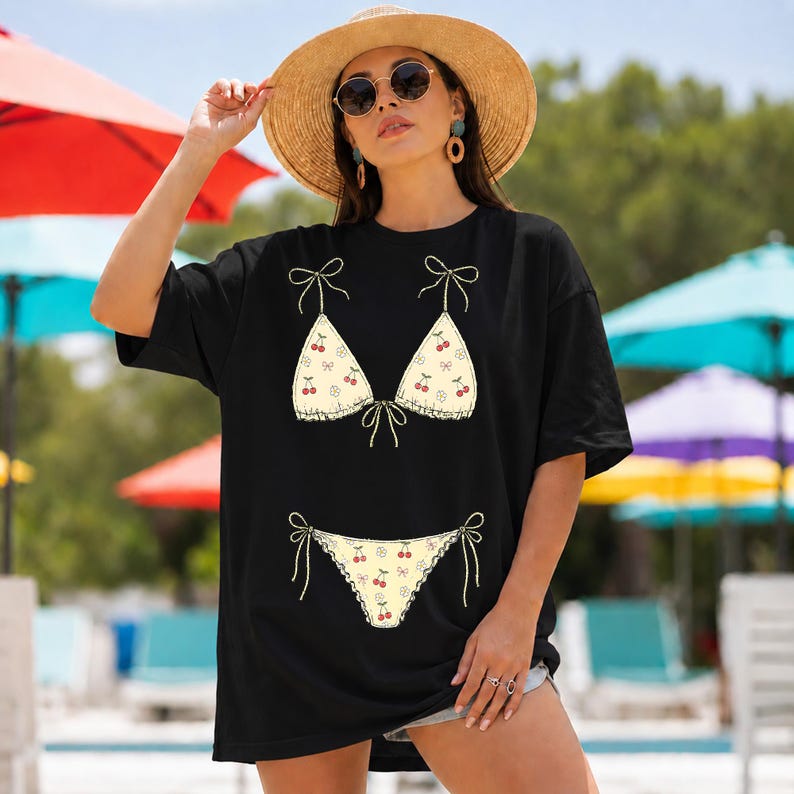 以下が含まれることがあります： ビキニのデザインが施された黒いTシャツ。ビキニのトップスとボトムは、さくらんぼの模様とレースのトリムが付いた薄い黄色です。シャツはゆったりとしたフィット感で、半袖です。モデルはサングラスと麦わら帽子を着用しています。