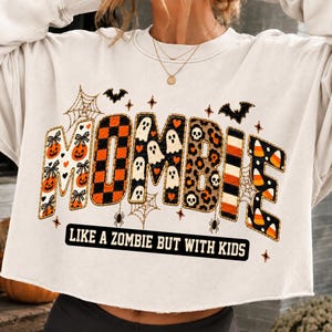 May include: A cream-colored cropped sweatshirt featuring the text MOMBIE in large, patterned letters filled with pumpkins, orange and black plaid, ghosts, leopard print, stripes, and candy corn. Below, the text reads LIKE A ZOMBIE BUT WITH KIDS.
