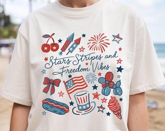 May include: Cream-colored t-shirt featuring a patriotic design with the text "Stars Stripes and Freedom Vibes." The graphic includes cherries, fireworks, an American flag, a hot dog, and stars in red, white, and blue.