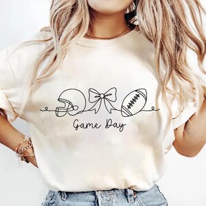 May include: Cream-colored sweatshirt with a black line drawing of a football helmet, bow, and football. The words "Game Day" are written in a script font below the drawing. The sweatshirt is a casual style.