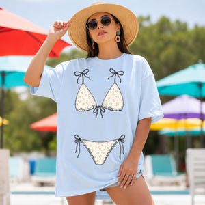 以下が含まれることがあります： ビキニのイラストが描かれた、水色のオーバーサイズのTシャツ。ビキニのトップスとボトムはクリーム色で、花柄と黒いリボンのディテールが施されています。サングラスと麦わら帽子をかぶった人が着ています。