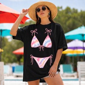 以下が含まれることがあります： パステルカラーのタイダイ柄ビキニデザインが特徴の黒いTシャツ。グラフィックには、リボンディテール付きのビキニトップとボトムが含まれています。このシャツは、夏のカジュアルなオーバーサイズスタイルです。