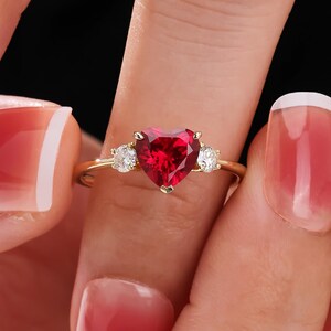 Könnte beinhalten: Ein goldener Ring mit einem herzförmigen, leuchtend roten Edelstein als Mittelpunkt, flankiert von zwei kleineren, runden, klaren Edelsteinen. Der Ring wird zwischen Fingern gehalten, um sein Design und seine Farbe hervorzuheben.