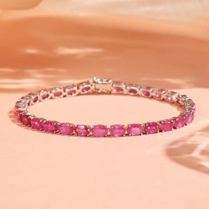 Könnte beinhalten: Ein silbernes Armband mit einer Reihe von oval geschliffenen, leuchtend rosa Edelsteinen. Die Edelsteine sind eng aneinander gesetzt und bilden eine durchgehende Farblinie. Das Armband wird auf einer zartrosa Oberfläche mit dezenten Schatten präsentiert.