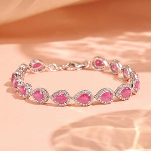 Pode incluir: Uma pulseira prateada com pedras preciosas rosas em forma de gota, cada uma rodeada por pequenas pedras transparentes. A pulseira é exibida sobre um fundo rosa suave, destacando o seu design elegante.