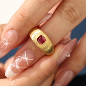 May include: A gold ring with a brushed finish, featuring a square, deep red gemstone at the center. The ring is set against a soft background, highlighting the rich color of the stone and the warm tone of the metal.
