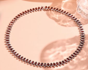 Puede incluir: Un collar de plata con una serie de piedras preciosas rojas oscuras talladas en forma de marquesa. Las piedras están engastadas juntas, formando una línea continua. El collar se exhibe sobre una superficie rosa claro.