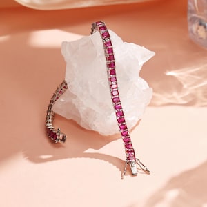 May include: A silver bracelet set with rectangular, ruby-red gemstones. The bracelet is presented on a white, textured surface against a soft pink backdrop. The clasp is visible, and the gemstones are evenly spaced.
