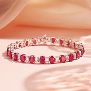 Op de afbeelding: Een zilveren armband met afwisselend ovale rode edelstenen en rechthoekige heldere edelstenen. De armband is geplaatst tegen een zachte roze achtergrond. De sluiting is zichtbaar. Een luxueus en elegant sieraad.