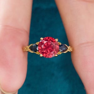 May include: A gold ring with a large, round, red gemstone at the centre, and two dark blue, pear-shaped stones either side. The band has an ornate, textured design.