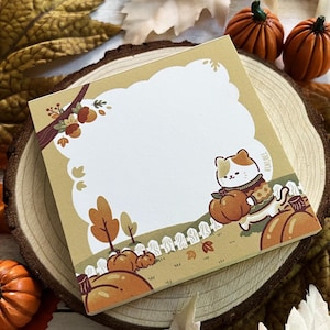 Peut inclure: Un bloc-notes carré avec une illustration sur le thème de l'automne représentant un chat portant une citrouille. Le bloc-notes est entouré de feuilles d'automne décoratives et de petites citrouilles. Le bloc-notes a un espace vide pour les notes.