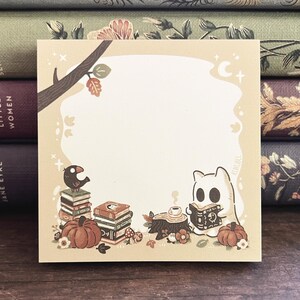 May include: Square notepad with a whimsical illustration of a ghost reading, pumpkins, and a bird on books. The notepad has a light beige background with a tree branch and autumn leaves. Ideal for notes.