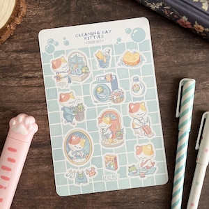Peut inclure: Une planche d'autocollants intitulée "Cleaning Day Kitties" avec des illustrations de chats faisant le ménage. La planche a un fond quadrillé bleu clair et comprend des autocollants d'une machine à laver, de produits de nettoyage et de chats. Deux stylos et un porte-stylo rose en forme de patte sont également visibles.