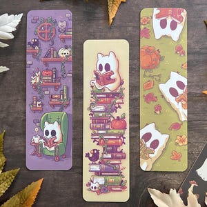 May include: Three Halloween-themed bookmarks. One shows a ghost reading on books, another has a ghost reading in a green chair, and the third features ghosts and pumpkins on a green background. The bookmarks are rectangular in shape.
