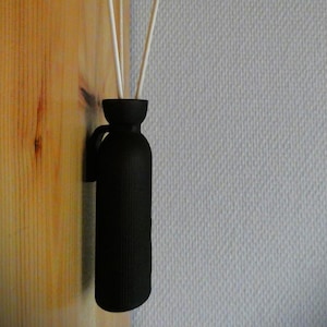 Puede incluir: Un difusor negro texturizado con asa, que contiene varillas blancas. La botella está montada sobre una superficie de madera, con una pared gris claro en el fondo. El difusor está diseñado para la fragancia del hogar.