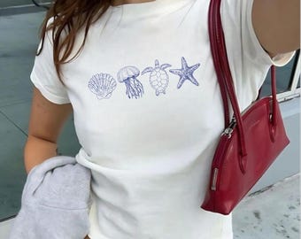 May include: White t-shirt with blue sea creature graphics, including a seashell, jellyfish, turtle, and starfish. A red handbag with a zipper closure is visible. The shirt is short-sleeved.