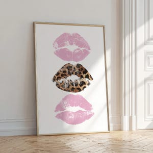 May include: A framed print featuring three lip prints. The top and bottom prints are a soft pink, while the middle print has a leopard print design. The artwork is displayed on a white background.