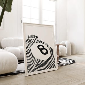 May include: A large, white framed print features a black and white zebra-striped design incorporating a black number eight billiard ball. The text 'Lucky' is written in a stylized black font above the ball. The print is leaning against a white wall, partially visible in the image. The print's overall theme is modern and minimalist, suitable for home decor.