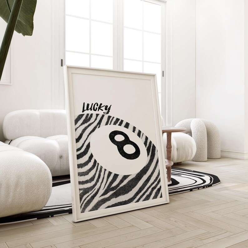 May include: A large, white framed print features a black and white zebra-striped design incorporating a black number eight billiard ball. The text 'Lucky' is written in a stylized black font above the ball. The print is leaning against a white wall, partially visible in the image. The print's overall theme is modern and minimalist, suitable for home decor.