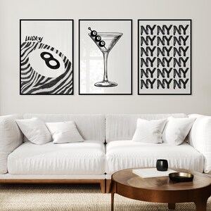 May include: Three black and white wall art prints. The left print features a zebra-striped design with a pool ball and the word 'Lucky'. The center print shows a martini glass with olives. The right print displays a repeating pattern of the letters 'NYNYNY'. The prints are displayed above a white couch and a wooden coffee table.
