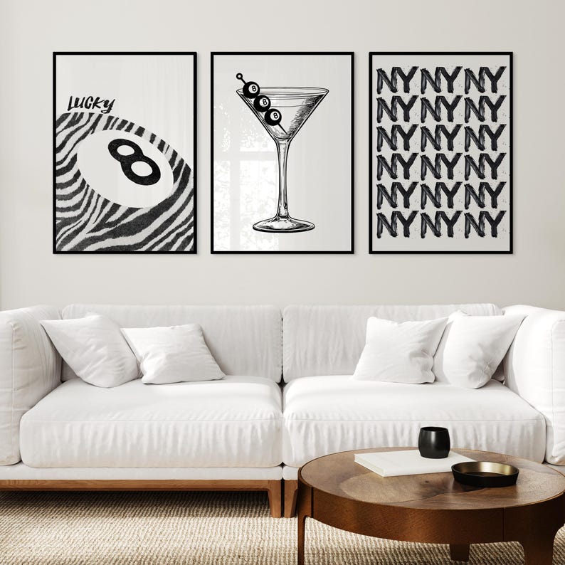 May include: Three black and white wall art prints. The left print features a zebra-striped design with a pool ball and the word 'Lucky'. The center print shows a martini glass with olives. The right print displays a repeating pattern of the letters 'NYNYNY'. The prints are displayed above a white couch and a wooden coffee table.