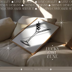 May include: A digital download of a minimalist black and white illustration of a martini glass with three olives. The text 'LUCKY GIRL LUXE' is displayed below the illustration. The image is displayed on a beige couch. The text at the top reads 'QUALITY LUXE QUALITY LUXE QUALITY LUXE' and 'TRENDY DESIGNS TRENDY DESIGNS TRENDY DESIGNS'. The bottom text reads 'DIGITAL DOWNLOAD'.