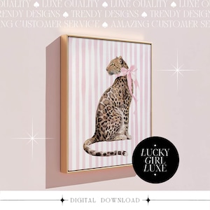 Op de afbeelding: Een ingelijste digitale print met een luipaard met een roze strik, tegen een achtergrond van roze en witte verticale strepen. Het kunstwerk zit in een gouden lijst, met de tekst "LUCKY GIRL LUXE" op een zwarte cirkel. Onderaan staat "DIGITAL DOWNLOAD".