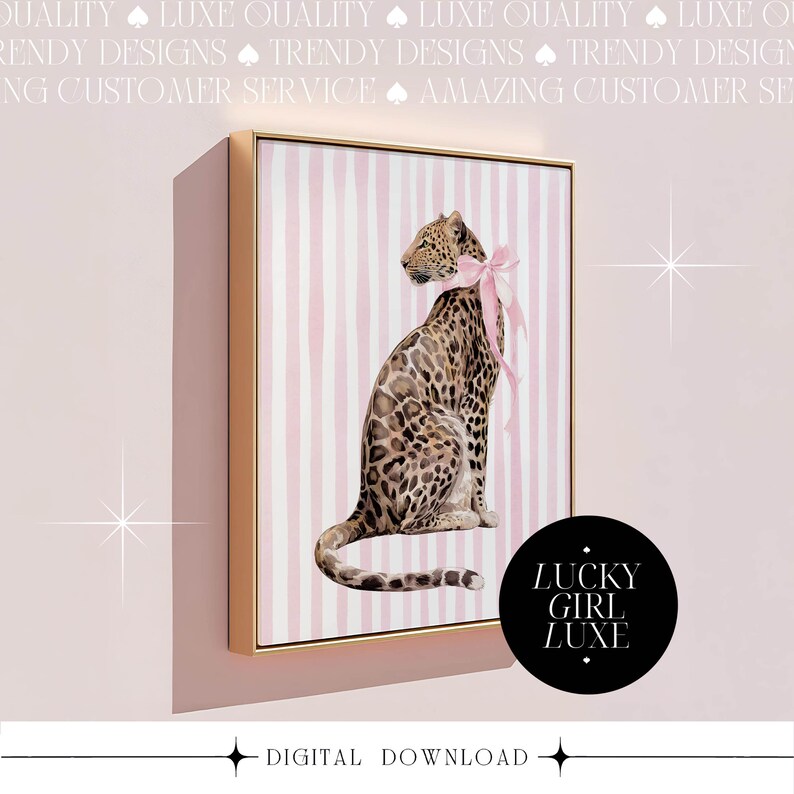 Op de afbeelding: Een ingelijste digitale print met een luipaard met een roze strik, tegen een achtergrond van roze en witte verticale strepen. Het kunstwerk zit in een gouden lijst, met de tekst "LUCKY GIRL LUXE" op een zwarte cirkel. Onderaan staat "DIGITAL DOWNLOAD".