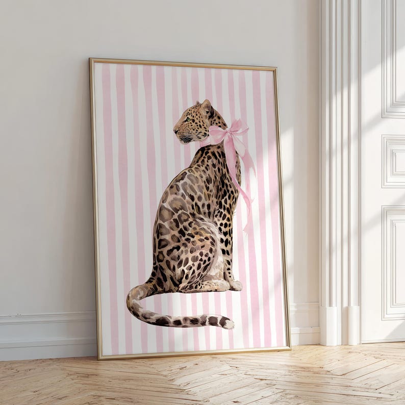 Op de afbeelding: Een ingelijste kunstdruk met een aquarel illustratie van een luipaard met een roze strik, tegen een roze en wit gestreepte achtergrond. Het kunstwerk wordt gepresenteerd in een gouden lijst, wat een vleugje elegantie toevoegt.