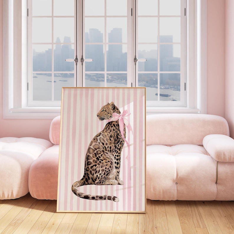 Op de afbeelding: Een ingelijste kunstdruk met een luipaard met een roze strik om zijn nek, tegen een achtergrond van verticale roze en witte strepen. De luipaard heeft een gevlekte vacht en zit rechtop. De lijst is goudkleurig.