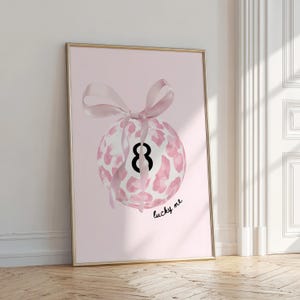 Puede incluir: Una impresión artística enmarcada con un fondo rosa, que representa una bola 8 blanca y rosa con estampado de leopardo, atada con una cinta rosa. El número 8 es negro. Las palabras "lucky me" están escritas en escritura negra.
