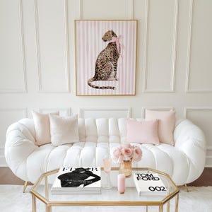 Op de afbeelding: Een ingelijste luipaardprint met een roze strik, boven een witte bank met roze kussens. Een glazen salontafel toont boeken, bloemen en een kaars. De kamer heeft een moderne, elegante uitstraling.