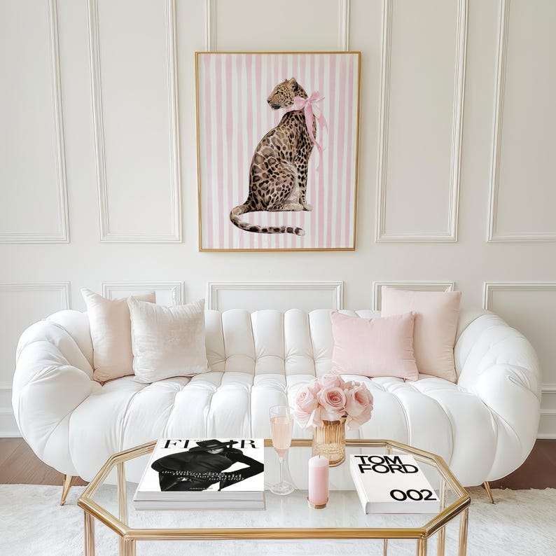 Op de afbeelding: Een ingelijste luipaardprint met een roze strik, boven een witte bank met roze kussens. Een glazen salontafel toont boeken, bloemen en een kaars. De kamer heeft een moderne, elegante uitstraling.
