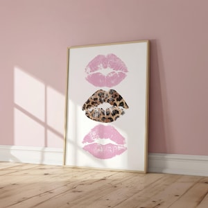 May include: Framed art print featuring three lip prints. Two pink lip prints flank a central lip print with a leopard print pattern. The artwork is displayed on a white background within a light wood frame, leaning against a pink wall.