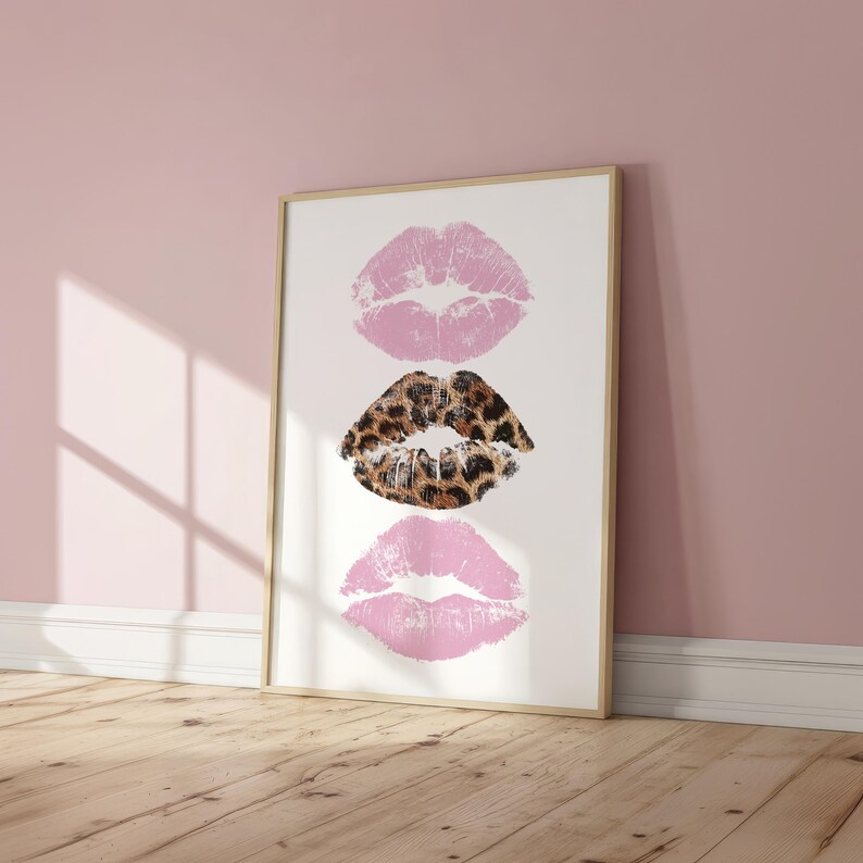 May include: Framed art print featuring three lip prints. Two pink lip prints flank a central lip print with a leopard print pattern. The artwork is displayed on a white background within a light wood frame, leaning against a pink wall.