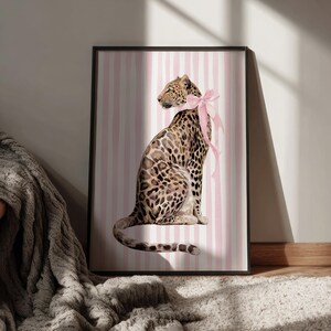 Op de afbeelding: Een ingelijste kunstdruk met een aquarelillustratie van een luipaard met een roze strik. De luipaard staat tegen een achtergrond van roze en witte verticale strepen. Het kunstwerk wordt gepresenteerd in een zwart frame.