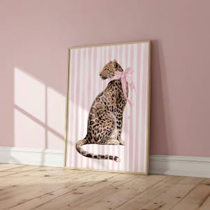 Op de afbeelding: Een ingelijste kunstdruk met een illustratie van een luipaard met een roze strik, tegen een achtergrond van roze en witte strepen. De lijst heeft een lichte houtkleur. Geschikt voor interieurdecoratie.