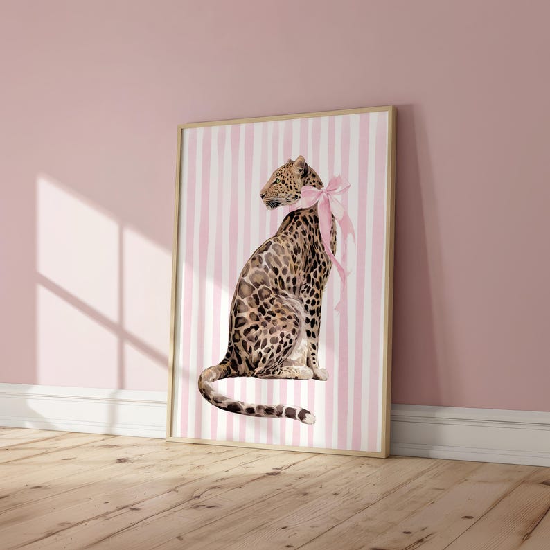 Op de afbeelding: Een ingelijste kunstdruk met een illustratie van een luipaard met een roze strik, tegen een achtergrond van roze en witte strepen. De lijst heeft een lichte houtkleur. Geschikt voor interieurdecoratie.