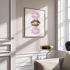 May include: Framed art print featuring three lip prints. Two pink lip prints flank a central leopard print lip print. The artwork is displayed in a gold frame against a white background, adding a touch of glamour to any space.