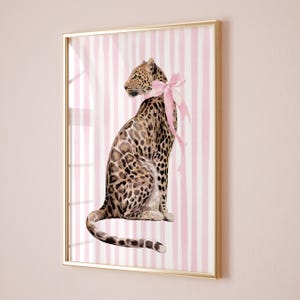 Puede incluir: Lámina artística enmarcada con una ilustración de acuarela de un leopardo con un lazo rosa alrededor del cuello. El leopardo se encuentra sobre un fondo de rayas verticales rosas y blancas, todo dentro de un marco dorado. La obra tiene una estética caprichosa y elegante.