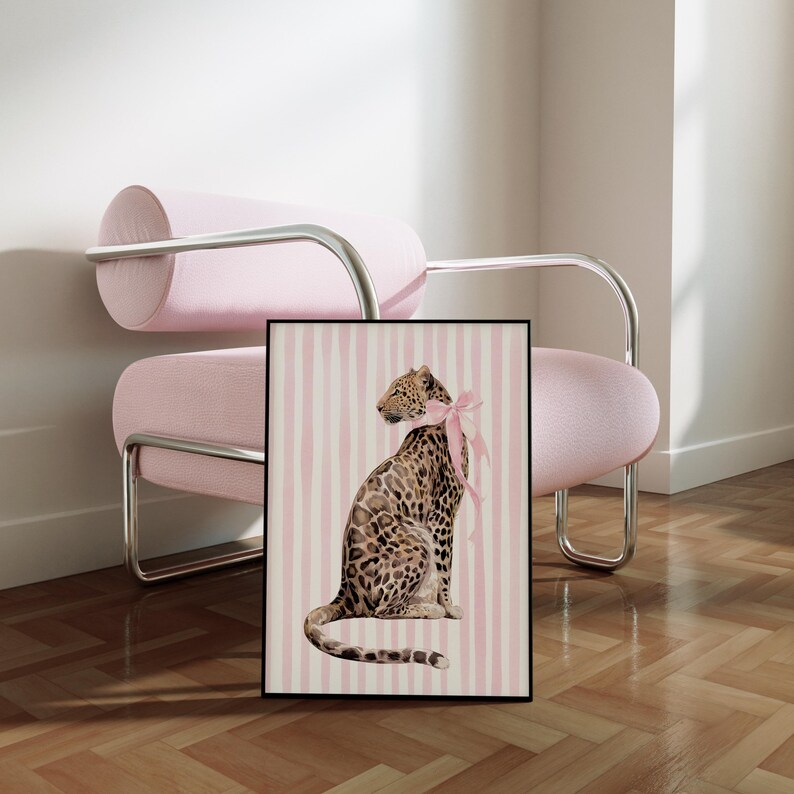 Op de afbeelding: Een ingelijste kunstdruk met een luipaard met een roze strik, tegen een roze en wit gestreepte achtergrond. De luipaard heeft een gevlekte vacht. De print zit in een zwart frame. De afbeelding wordt weergegeven in een kamer met een roze stoel.