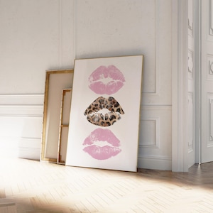 May include: A framed art print featuring three lip imprints. The top and bottom lips are a soft pink, while the middle lip print has a leopard print design. The artwork is displayed against a white background, leaning against a wall.