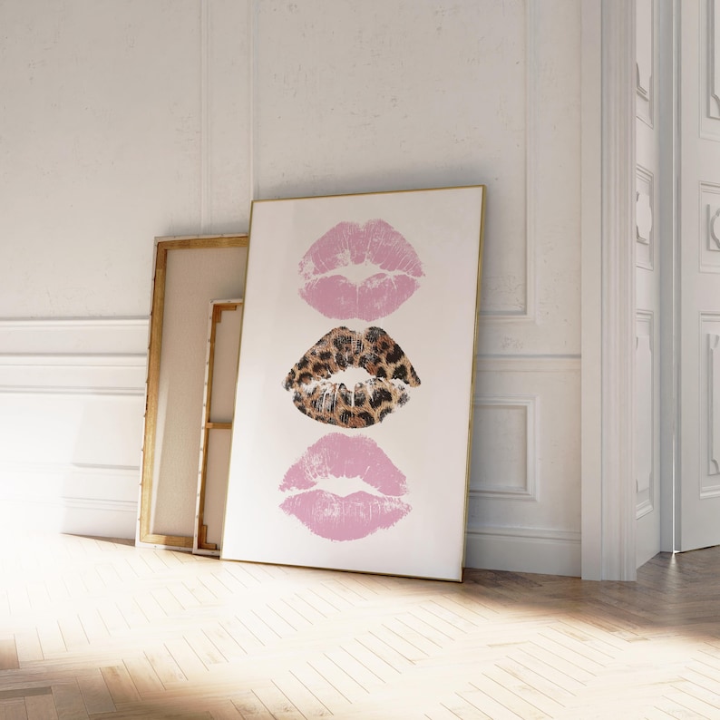 May include: A framed art print featuring three lip imprints. The top and bottom lips are a soft pink, while the middle lip print has a leopard print design. The artwork is displayed against a white background, leaning against a wall.