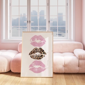 May include: A framed art print featuring three lip prints. The top and bottom prints are light pink, while the middle print has a leopard print design. The artwork is displayed in a room with a pink sofa and a window.