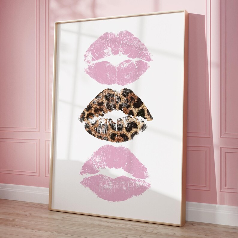 May include: A framed art print featuring three lip prints. Two are light pink, and the middle one has a leopard print pattern. The artwork is displayed on a white background, framed in a gold-colored frame, and leans against a pink wall.
