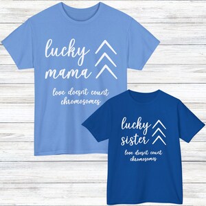 Op de afbeelding: Een lichtblauw t-shirt met witte tekst lucky mama en love doesn't count chromosomes met drie witte chevrons, en een koningsblauw t-shirt met witte tekst lucky sister en love doesn't count chromosomes met drie witte chevrons.