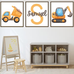 May include: Three framed prints featuring a yellow construction truck, a yellow letter S, and a yellow excavator. The letter S is surrounded by the name Samuel. The prints are hanging on a white wall above a grey shelf with baskets and toys.