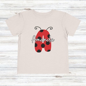 May include: A light beige toddler t-shirt featuring a red ladybug-shaped letter "A" with black spots. The name "Amanda" is written in cursive across the ladybug design. The shirt has short sleeves and a crew neck.